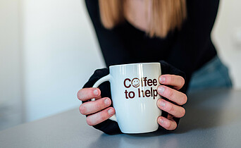 Ein junge Mädchen mit langen hellbraunen Haaren hält eine Coffee to help-Tasse in der Hand.