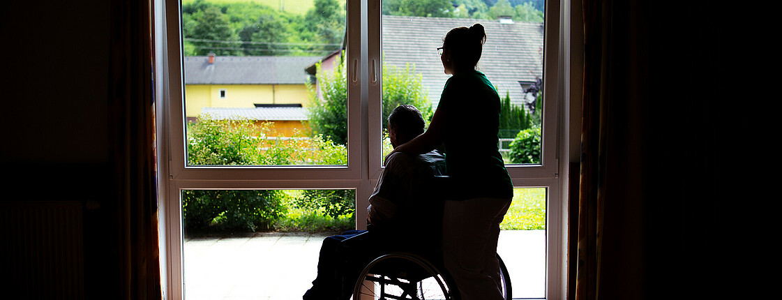 Eine junge Dame steht mit einem Mann im Rollstuhl vor einem beleuchteten Fenster, deshalb sieht man nur die Umrisse der Beiden.