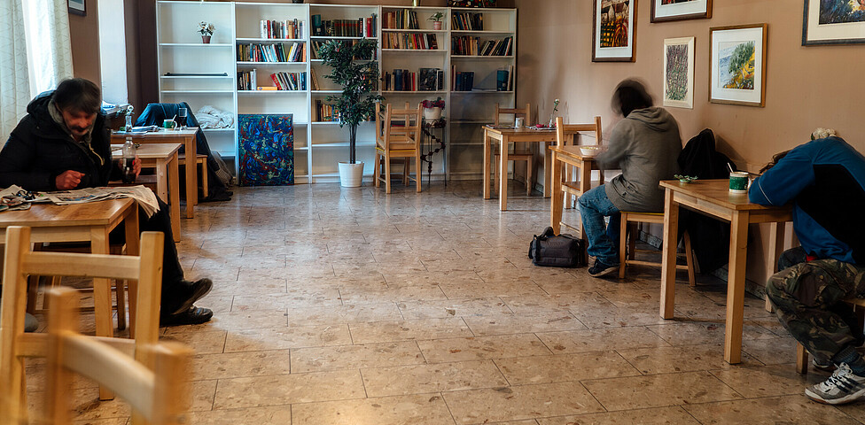 Der Aufenthaltsraum der Wohnungslosentagesstätte "Eggerheim" links und rechts von der Wand stehen Tische, auf den auch Besucher*innen sitzen. Hinten an der Wand des Raumes steht ein großes Regel gefüllt mit Büchern.