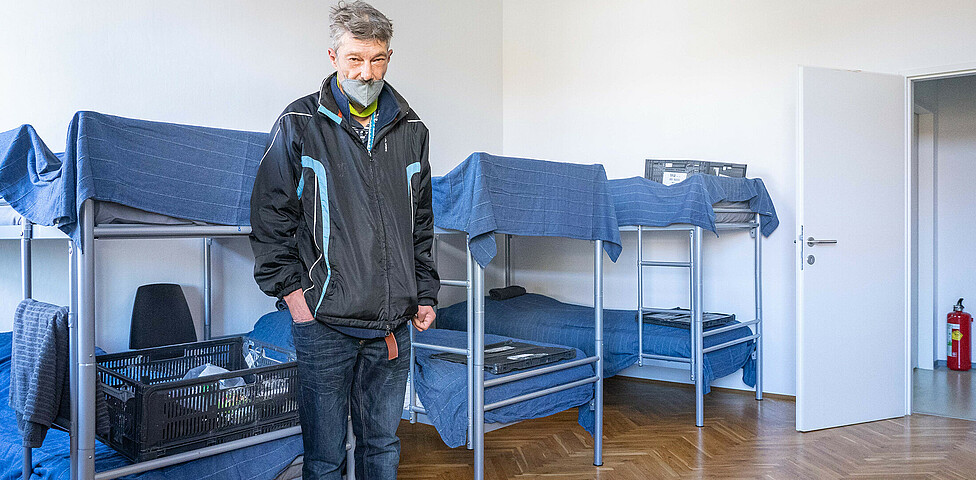 Mehrere Hochbetten stehen in der Notschlafstelle vor einem steht ein wohnungsloser Mann und seine Habseligkeiten liegen auf einem der Betten.