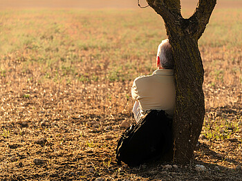 Ein älterer Herr sitzt einsam an den Stamm eines Baum gelehnt auf einer Wiese und sieht in die Ferne.