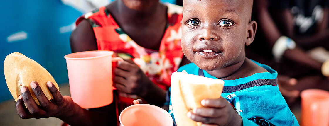 Zwei Kinder in Uganda sitzen in der Schule und haben ein frischgebackenes Brot sowie bunte Trinkbecher in der Hand.
