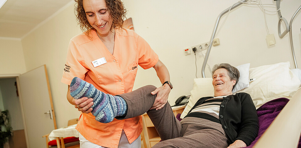 Eine Pflegekraft hilft einer Bewohnerin, die im Bett liegt mit Bewegungsübungen ihres Beines.