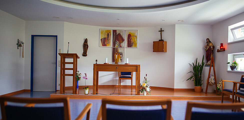 Ein paar Stühle stehen vor einem Altar mit Blumen und Kerzen, Rednerpult, 2 Kreuze, Christusfigur und 4 aufgehängte Bilder.