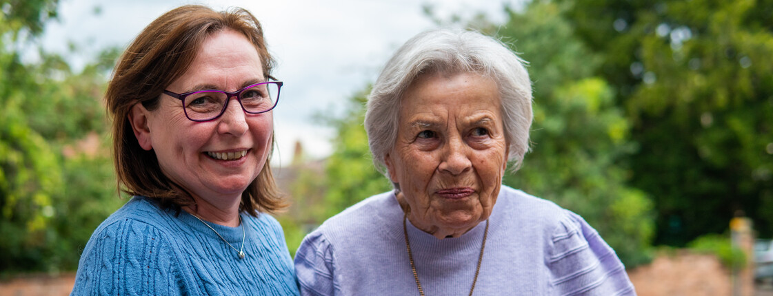 Eine Frau mittleren Alters mit Brille stützt eine Senorin, die eine Halskette trägt, beim Gehen. Im Hintergrund sind grüne Bäume.