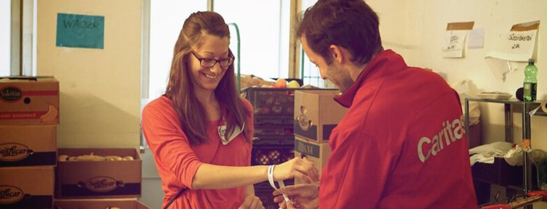 Ein Mitarbeiter der Caritas bindet einer freiwilligen Helferin in einer Lebensmittel-Ausgabestelle der Caritas ein Armband um als Dankeschön für ihre Mithilfe. Im Hintergrund sind Kartons mit diversen Lebensmitteln zu sehen.