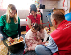 Kinder sitzen mit Betreuungspersonen auf Teppich und spielen Lernspiele, unter den Personen auch Projektreferentin Bettina Zangl-Jagiello bei ihrem Projektbesuch