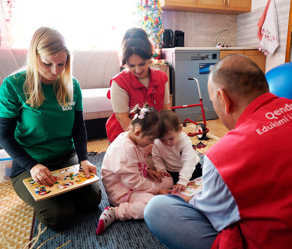Kinder sitzen mit Betreuungspersonen auf Teppich und spielen Lernspiele, unter den Personen auch Projektreferentin Bettina Zangl-Jagiello bei ihrem Projektbesuch