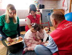 Kinder sitzen mit Betreuungspersonen auf Teppich und spielen Lernspiele, unter den Personen auch Projektreferentin Bettina Zangl-Jagiello bei ihrem Projektbesuch