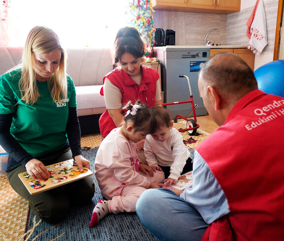 Kinder sitzen mit Betreuungspersonen auf Teppich und spielen Lernspiele, unter den Personen auch Projektreferentin Bettina Zangl-Jagiello bei ihrem Projektbesuch