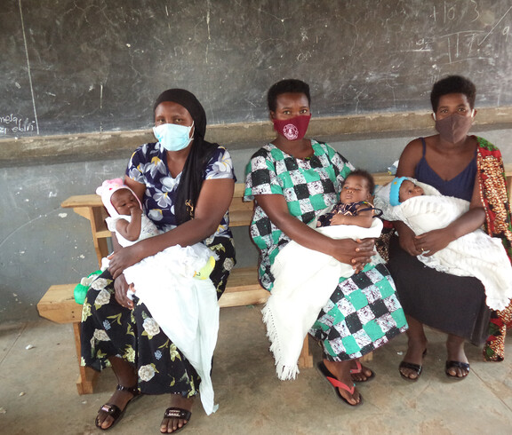 Drei Mütter sitzen mit ihren Kindern im Arm auf einer Bank und warten.