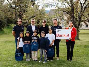 Acht Kinder mit Rucksäcken und sechs Erwachsene bei Caritas-Übergabeaktion im Grünen, Schild mit „Danke!“