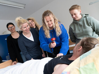Eine Schülerin misst bei einer Mitschülerin den Blutdruck, die in einem Krankenbett liegt, die Lehrerin und Mitschüler*innen beobachten sie dabei.