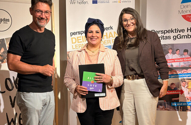 Drei Personen stehen vor einer Wand mit Plakaten, eine Frau hält Broschüren.