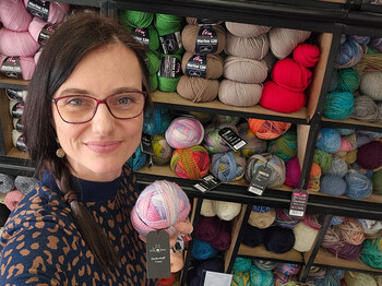 Person hält Wollknäuel in Stoffgeschäft vor Regalen mit Wolle beim Mützchen-Stricken für Caritas und Innocent.