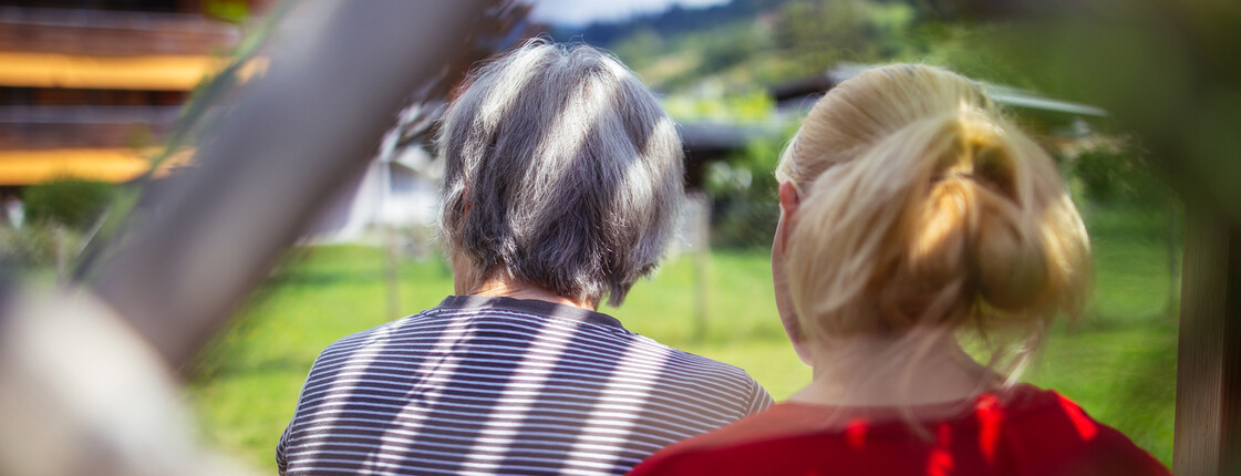 Eine ältere Dame und eine junge Frau sitzen nebeneinander in einer Laube, beide sieht man nur von hinten.