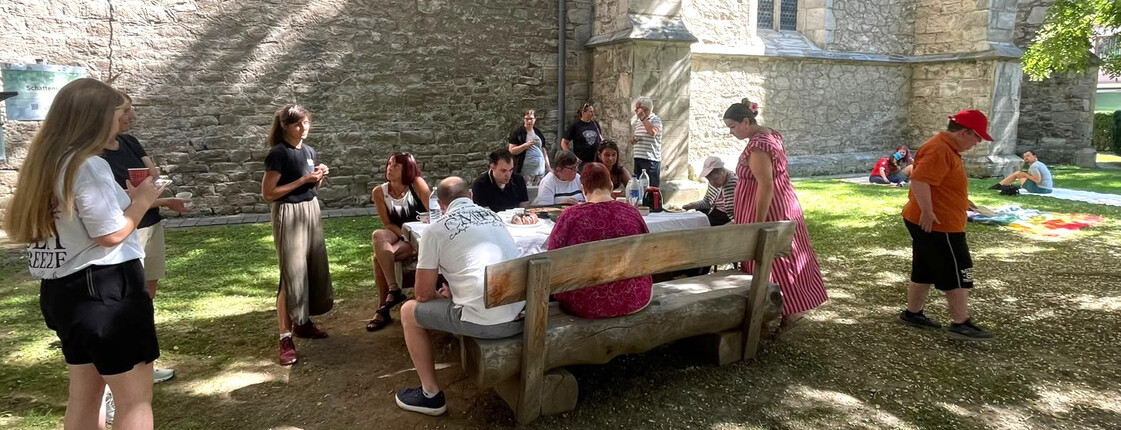 Menschen sitzen und stehen an einem Tisch im Schatten neben einer Pfarrkirche und kommen miteinander ins Gespräch.