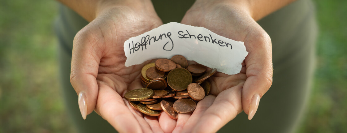 Zwei Hände formen eine Schüssel, in dieser liegen Geldmünzen und ein Zettel mit dem Schriftzug "Hoffnung schenken".