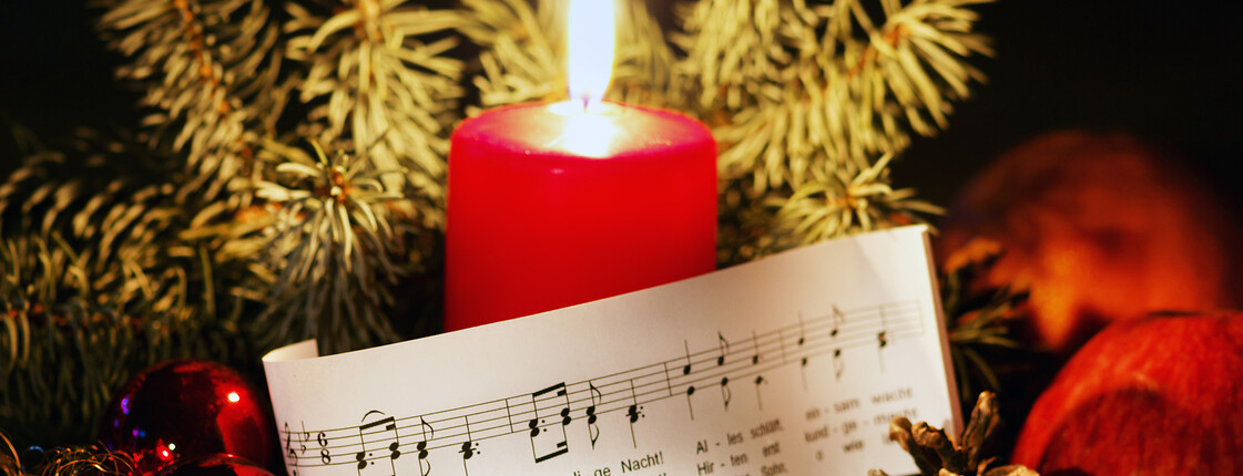 Rote Kerze zwischen Tannenzweigen, Christbaumkugeln und Notenblatt mit „Stille Nacht“ als Motiv der Weihnachtsandacht.