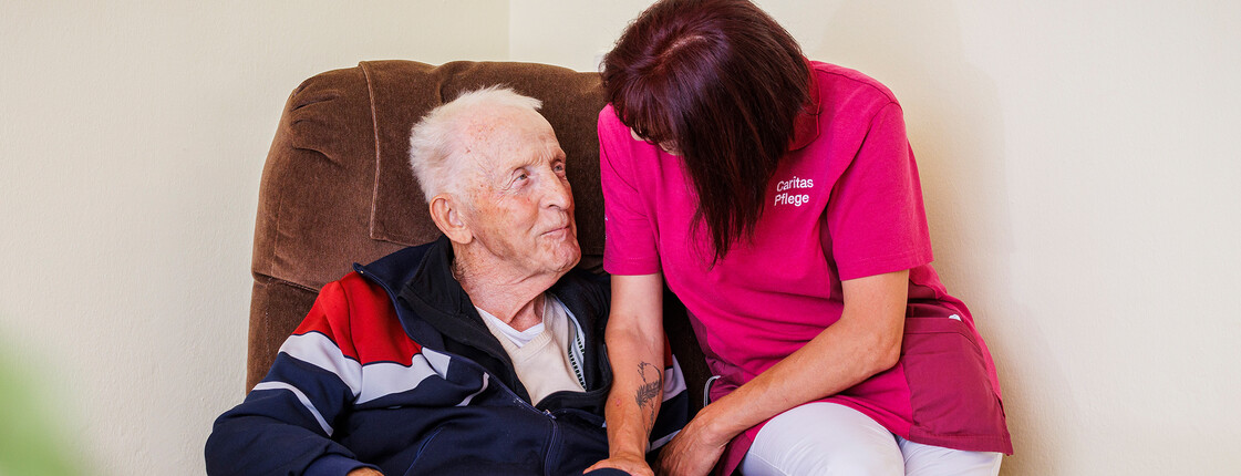 Pflegekraft sitzt neben einem älteren Herrn auf dem Sessel.