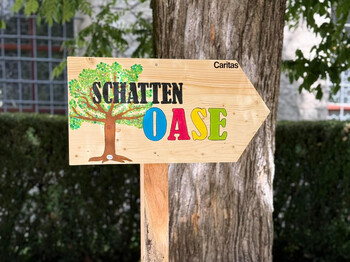 Holzschild mit der Aufschrift „Schatten Oase“ und Caritas-Logo vor einem Baum im Freien.
