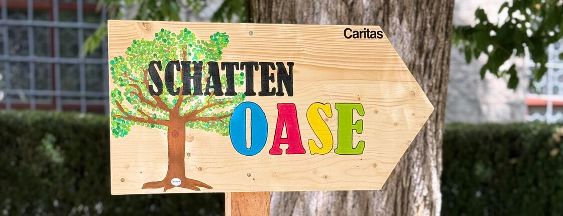 Holzschild mit der Aufschrift „Schatten Oase“ und Caritas-Logo vor einem Baum im Freien.