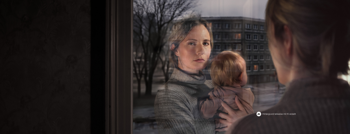 Eine Frau mit Kind am Arm schaut aus dem Fenster. Ihr Gesicht spiegelt sich im Fensterglas wider. Vor dem Fenster ist eine Wohngegend erkennbar.