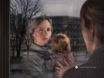 Eine Frau mit Kind am Arm schaut aus dem Fenster. Ihr Gesicht spiegelt sich im Fensterglas wider. Vor dem Fenster ist eine Wohngegend erkennbar.