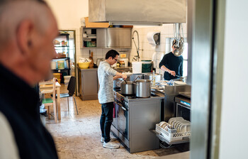 Ein Besucher unseres Eggerheims im Bild rechts und unscharf dargestellt, sieht in die Küche der Tagesstätte in der gerade zwei Zivildiener das Mittagessen vorbereiten.