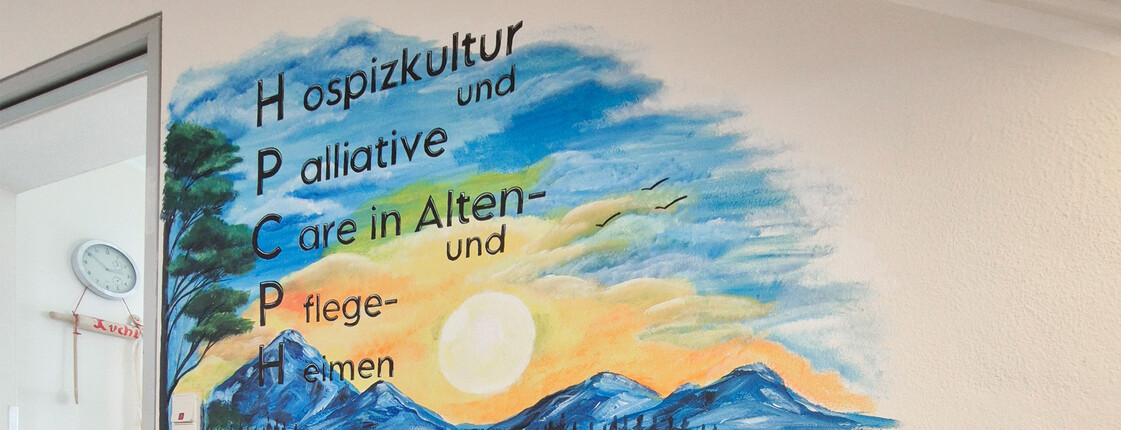 Flur Pflegewohnhaus mit Wandgemälde aus Bergen und Sonnenuntergang, Text „Hospizkultur und Palliative Care“, Treppe im Vordergrund.