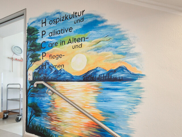 Flur Pflegewohnhaus mit Wandgemälde aus Bergen und Sonnenuntergang, Text „Hospizkultur und Palliative Care“, Treppe im Vordergrund.