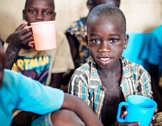 Ein Kind mit einer Zahnlücke vorne, lächelt in die Kamera, während er einen blauen Becher in der Hand hält. Um ihn herum sind seine Klassenkameraden, die etwas aus dem Bechern trinken.