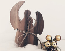 Zwei Engel aus Holz sind auf Watte und mit goldenen Kugeln verziert vor einem weißen Hintergrund drapiert.