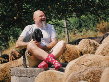 "Shoafbauer" Thomas Koch sitzt zwischen Schafen im Freien und hält ein Lamm auf dem Schoß; Szene vermittelt Nähe zu Schafhaltung und Handwerk.