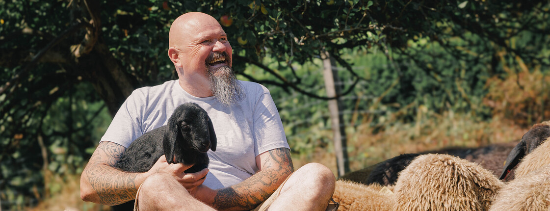 "Shoafbauer" Thomas Koch sitzt zwischen Schafen im Freien und hält ein Lamm auf dem Schoß; Szene vermittelt Nähe zu Schafhaltung und Handwerk.