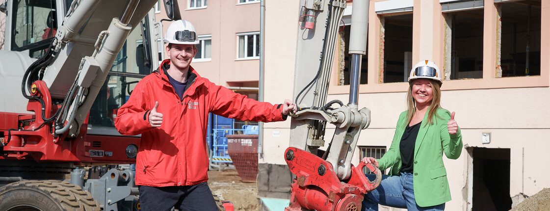 Zwei Personen mit Helmen stehen vor Bagger auf Baustelle und zeigen Daumen hoch im Kontext des Caritas Social Deals.