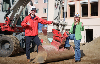 Zwei Personen mit Helmen stehen vor Bagger auf Baustelle und zeigen Daumen hoch im Kontext des Caritas Social Deals.