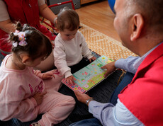 Betreuungsperson zeigt zwei Kindern ein buntes Sprachbuch auf dem Boden.