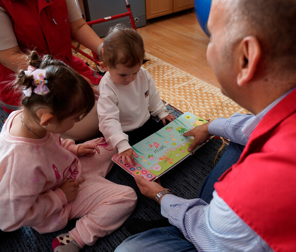 Betreuungsperson zeigt zwei Kindern ein buntes Sprachbuch auf dem Boden.