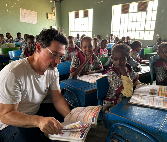 Schüler*innen sitzen im Klassenzimmer. Ernst Sandriesser sieht gemeinsam mit einer Schülerin in ein Schulbuch.