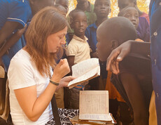 Natürlich — hier ist der überarbeitete Alternativtext als ausgewogener Mix beider Varianten:  > **Alternativtext:** > Die Bereichsleiterin Alexandra Blattnig-Rull der Auslandshilfe der Caritas Kärnten sitzt bei einem Projektbesuch und liest in einem Heft, während sie von Kindern der Schule umringt ist und mit ihnen spricht.