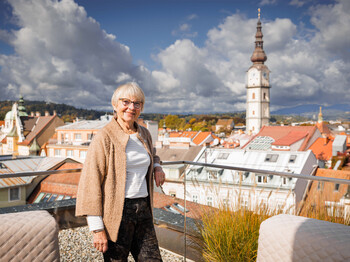 Martha Mann, Darlehensgeberin des Social Deals der Caritas Kärnten, steht auf einer Dachterrasse in Klagenfurt, im Hintergrund sind Dächer und ein Kirchturm zu sehen.