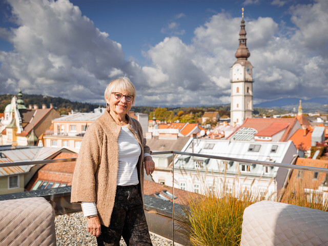 Martha Mann, Darlehensgeberin des Social Deals der Caritas Kärnten, steht auf einer Dachterrasse in Klagenfurt, im Hintergrund sind Dächer und ein Kirchturm zu sehen.