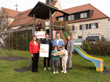 Vertreter*innen der Volksbank mit Caritas-Team und Kindern vor Rutsche im Garten eines Lerncafés, halten Schilder mit „Wir helfen“ und „Danke!“