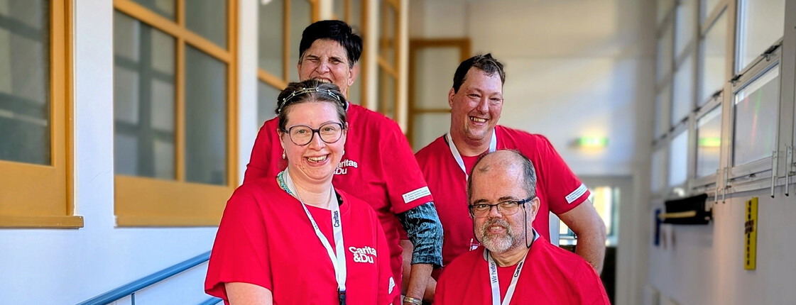 Vier Personen in roten Caritas-T-Shirts stehen gemeinsam im Flur und engagieren sich im inklusiven Projekt.