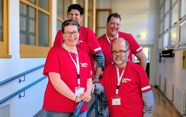 Vier Personen in roten Caritas-T-Shirts stehen gemeinsam im Flur und engagieren sich im inklusiven Projekt.
