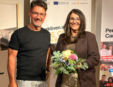 Caritasdirektor Ernst Sandriesser steht mit Projektleiterin Sabine Schnitzer, die einen Blumenstrauß hält, vor einem Caritas-Banner bei einer Projektveranstaltung. 