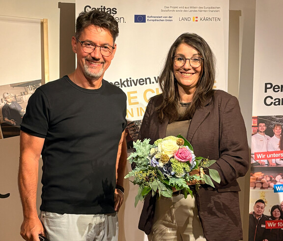 Caritasdirektor Ernst Sandriesser steht mit Projektleiterin Sabine Schnitzer, die einen Blumenstrauß hält, vor einem Caritas-Banner bei einer Projektveranstaltung. 