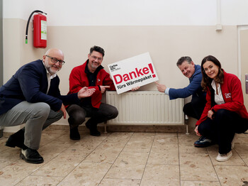 Vier Personen zeigen vor Heizkörper ein Schild „Danke fürs Wärmepaket“ im Eggerheim der Caritas.