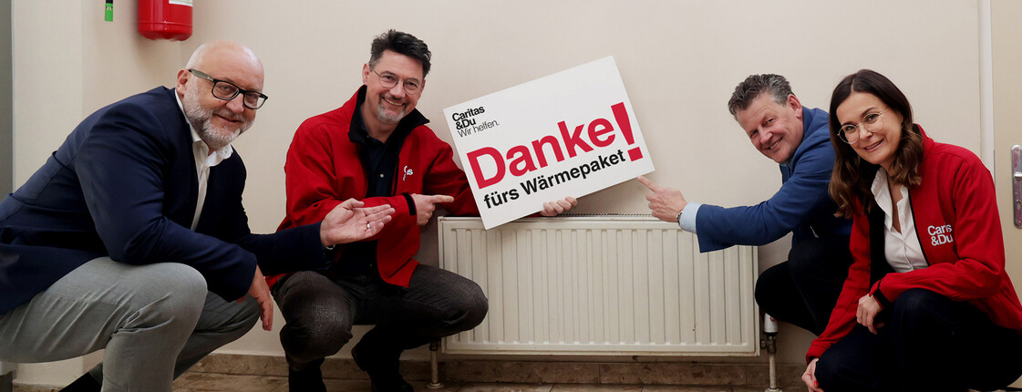 Vier Personen zeigen vor Heizkörper ein Schild „Danke fürs Wärmepaket“ im Eggerheim der Caritas.